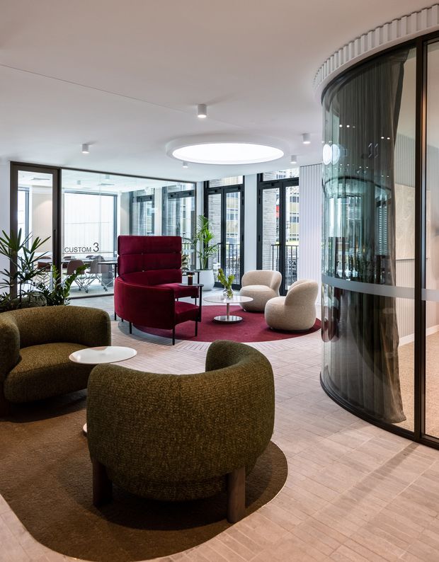 A refined contemporary commercial lounge or reception area with curved olive-green upholstered seating, a burgundy high-back chair, cream lounge chairs, circular tables, and layered circular rugs. The space has pale timber-look flooring, extensive black-framed glazing, a curved glass partition, ribbed wall detailing, abundant daylight, and a large circular ceiling light.