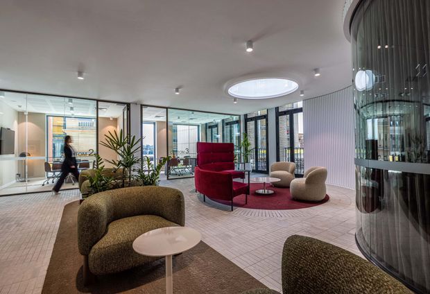 Contemporary commercial lounge or reception area with curved upholstered seating arranged around low round tables and a red circular rug. The space has a light tiled floor, white ceiling with recessed spotlights and a large oval skylight, glazed meeting rooms, black-framed glass partitions, vertical wall panelling, indoor planting, and a prominent curved glass feature enclosure.