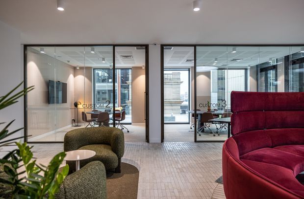 Contemporary commercial office interior with glazed meeting rooms labelled Custom 2 and Custom 3, open-plan workspaces, desks and chairs, and a foreground lounge area with rounded green and deep red upholstered seating. The space has a pale tiled floor, white ceiling with recessed downlights, dark-framed internal glazing, and a bright, professional atmosphere.