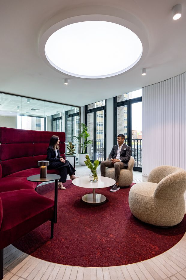 A contemporary commercial lounge or reception area with floor-to-ceiling glazing, dark-framed windows, a large circular skylight, and vertical white wall panelling. The space features a deep burgundy curved banquette and circular rug, sculptural cream boucle lounge chairs, round coffee tables, indoor plants, and two people seated in conversation. The palette is refined and high-contrast, combining burgundy, cream, white, black, and natural timber tones.