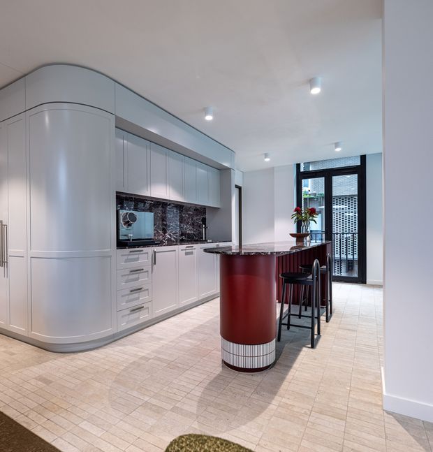 A refined contemporary kitchen with curved white built-in cabinetry, integrated appliances, dark marble-look splashback, pale tiled flooring, and a distinctive cylindrical burgundy-red island with a ribbed metal base. A compact peninsula with bar stools sits near a black-framed glazed door, with recessed ceiling lights and a small floral arrangement adding detail.