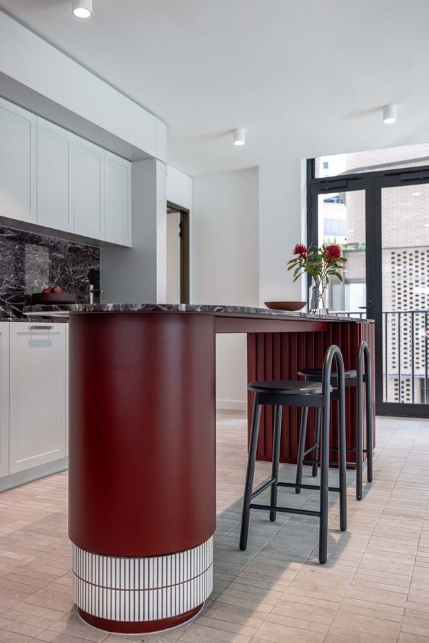 A bright contemporary kitchen centred on a large burgundy-red curved island with a dark veined stone benchtop and integrated vertical ribbed detailing. The island includes a rounded end with white slatted trim and a red base, paired with dark bar stools. White shaker-style cabinetry, a dark marble-look splashback, pale tiled flooring, recessed and surface-mounted lighting, and black-framed glazed doors complete the space.
