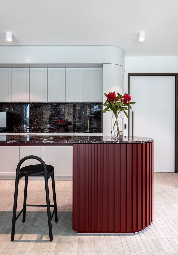A refined contemporary kitchen with pale paneled cabinetry, a dramatic dark veined stone backsplash and benchtop, and a rounded island featuring deep burgundy vertical fluted cabinetry. A black bar stool sits at the island, while a curved white bulkhead and concealed lighting add sculptural detail. The space has a light tiled floor and a framed doorway in the background.