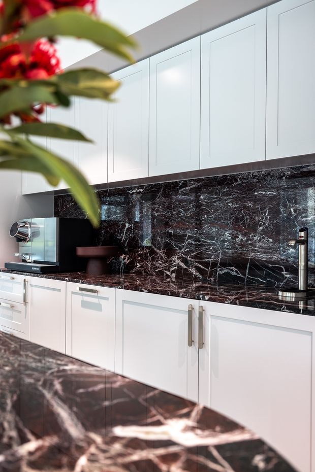 Close-up view of a refined contemporary kitchen with white framed cabinetry, dark richly veined marble benchtops and matching full-height splashback, brushed metal handles and tapware, and a small coffee station. A blurred leafy plant appears in the foreground.