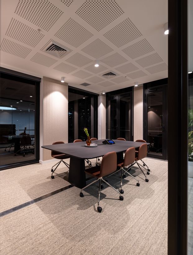 A contemporary commercial meeting room with a large dark conference table surrounded by brown upholstered swivel chairs. Floor-to-ceiling black-framed glazing encloses adjacent offices, while warm vertical wall panelling, recessed downlights, acoustic perforated ceiling panels, and a neutral carpet create a refined corporate interior.