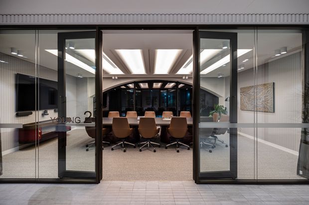 Contemporary commercial office interior viewed through glazed meeting-room doors. A large conference table with rolling chairs sits beneath a sculptural illuminated ceiling, with adjacent workspaces, a wall-mounted display, indoor plants, and framed artwork visible through the glass partitions.