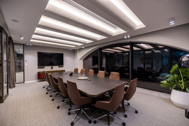 A contemporary boardroom with a large oval-rectangular conference table surrounded by tan upholstered swivel chairs. The room features a wall-mounted screen, integrated technology, grey carpet, dark-framed glazed partitions, and a sculptural recessed ceiling with linear lighting. A glazed adjoining space is visible beyond the arched opening.