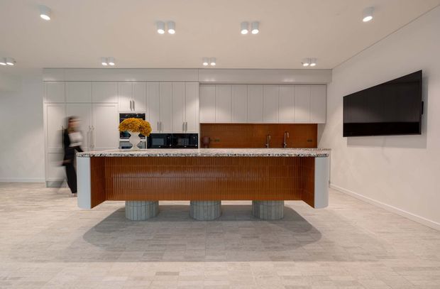 A bright, open-plan contemporary kitchen dominated by a large island with a ribbed timber front and stone benchtop. The kitchen features full-height white cabinetry, a warm timber splashback, integrated appliances, pendant-free recessed lighting, and sculptural cylindrical supports. A wall-mounted television is visible on the right.