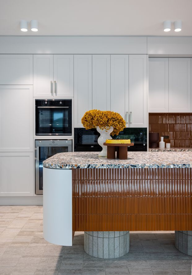 A refined contemporary kitchen featuring white Shaker-style cabinetry, integrated black and stainless-steel appliances, a dark splashback, and a large curved island. The island has a polished multicoloured stone benchtop, ribbed brown timber cabinetry, pale blue curved end panels, and cylindrical tiled supports. Decorative dried flowers and a bowl sit on the island, while a timber slatted feature is visible at the rear.