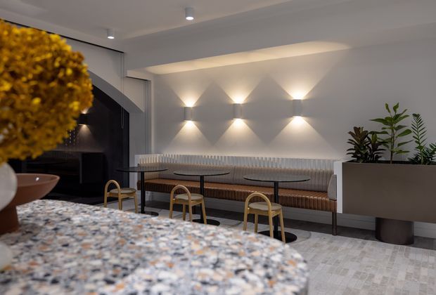 A refined contemporary hospitality-style interior with a long upholstered banquette, small round tables, and curved-back dining chairs arranged against a feature wall. Layered wall sconces create a geometric light pattern, while a speckled stone or terrazzo-like counter occupies the foreground. Large planters and sculptural floral decor add softness to the otherwise neutral, polished space.
