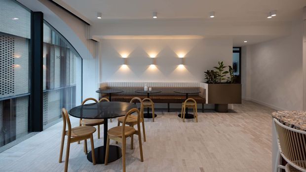 A refined contemporary communal lounge or dining area with round dark tables, curved timber chairs, a long upholstered banquette, and a large planter. The space features pale stone-look flooring, warm wall-wash lighting, neutral walls, and extensive curved glazing with patterned screening.