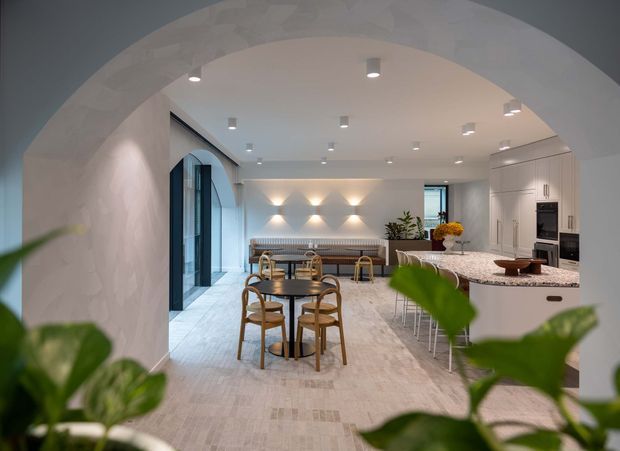 A refined open-plan kitchen and dining space viewed through a large rounded archway. The room features pale stone or plaster finishes, a light tiled floor, white built-in cabinetry, a large stone island with bar stools, a round dark dining table with timber chairs, arched glazing, and layered recessed and wall lighting. Green foliage in the foreground partially frames the scene.