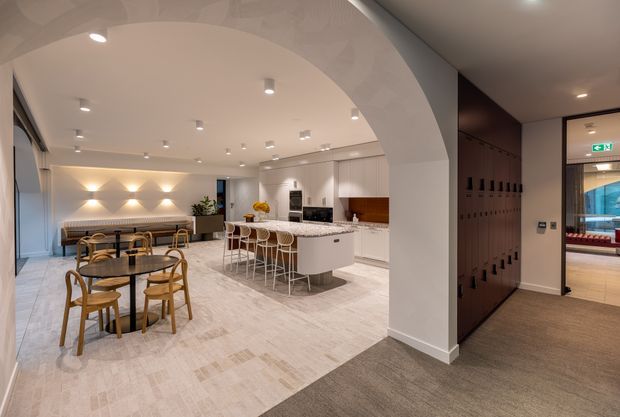 A refined open-plan hospitality or amenity space viewed through a broad arched opening. The room includes a white kitchen or refreshment island with bar stools, a dining area with round tables and timber chairs, built-in bench seating, soft geometric wall lighting, pale stone-look flooring, and a dark timber locker or cabinetry wall leading toward a glazed adjoining area.