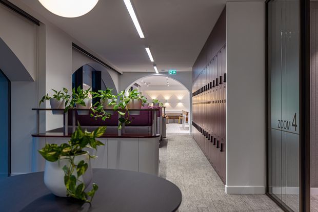 Contemporary commercial interior corridor with a reception or lounge zone, featuring burgundy upholstered seating, integrated planter dividers, extensive built-in locker or storage joinery, glazed rooms labelled “Zoom 4,” arched openings, linear ceiling lighting, and light grey carpet.