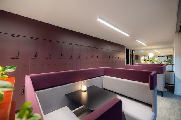 A contemporary commercial interior with a long wall of burgundy locker cabinetry, upholstered booth seating in burgundy and light grey, black tables, linear pendant lighting, and indoor plants. The space appears to be a communal breakout or lounge area, with glazed partitions and circulation areas visible beyond.