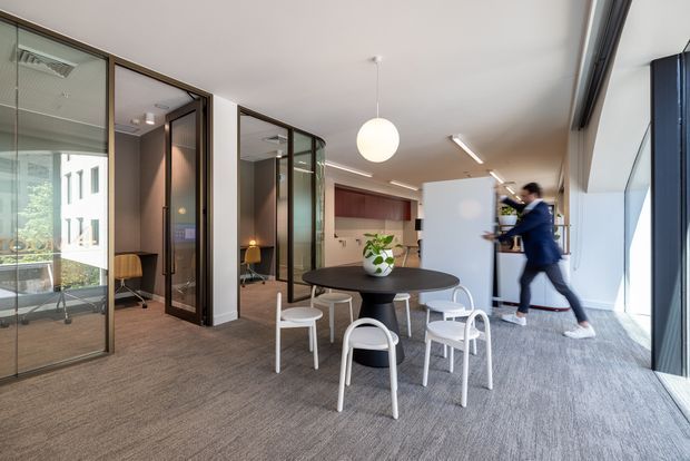 A bright contemporary open-plan workplace with a round black dining or meeting table and white chairs in the foreground. Glazed black-framed meeting rooms line the left side, while a kitchenette with dark timber cabinetry is visible at the rear. Full-height glazing on the right brings in abundant daylight, and a spherical pendant light hangs above the table. A blurred person is moving through the space.
