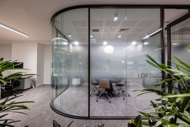 A contemporary commercial office interior featuring a curved, black-framed glass meeting room with frosted lower glazing, a conference table and rolling chairs inside, carpeted flooring, suspended linear lighting, acoustic ceiling panels, and indoor plants in the foreground. The word “LOFTUS” is visible on the meeting-room glass.