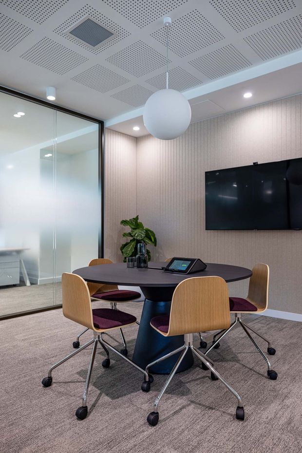 Contemporary commercial meeting room with a round dark conference table, four timber-shell swivel chairs with burgundy upholstery, wall-mounted display screen, and a video-conferencing console. The room features pale vertical acoustic wall panelling, grey carpet, a glazed partition, recessed lighting, and a perforated acoustic ceiling with a large spherical pendant light.