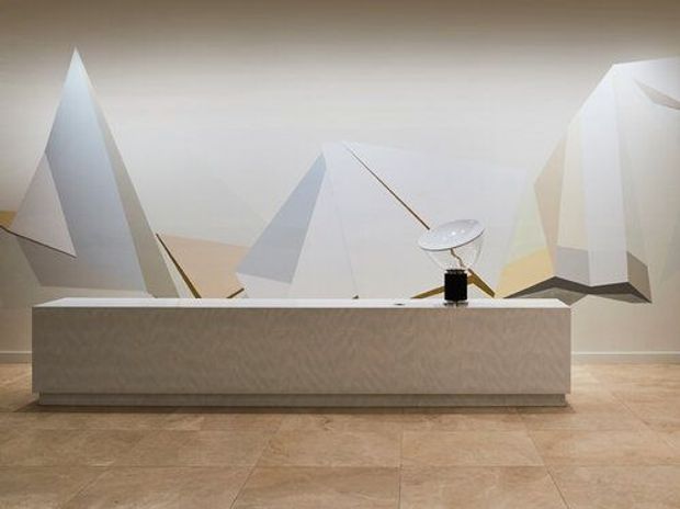 A minimalist reception or service counter with a long light-toned built-in front, set against a sculptural geometric wall installation composed of angular white and pale grey forms. The space has a pale tiled floor, neutral walls, and restrained contemporary lighting.