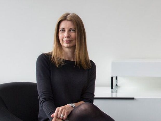 Portrait of a woman seated in a minimalist, predominantly white interior. A black upholstered chair and a simple white built-in architectural feature or shelving element are visible behind her.