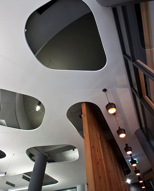 Upward-looking view of a contemporary interior with a sculptural white ceiling punctuated by several irregular rounded recessed openings. Dark cylindrical columns, exposed timber wall cladding, black-framed glazing, and a row of warm pendant lights create a dramatic architectural ceiling and circulation space.