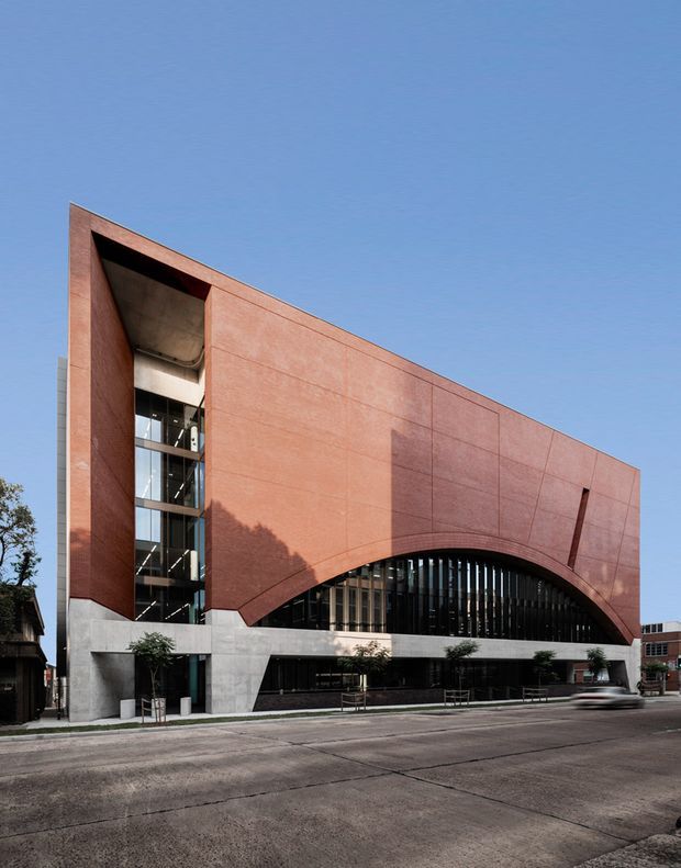 Street-facing view of a large contemporary institutional or commercial building. The facade has a strong geometric composition with a sloping terracotta-red panelled upper volume, a dramatic large arched opening filled with vertical dark glazing fins, exposed concrete at the base, and a tall glazed circulation or entrance slot at the left. The building sits in an urban streetscape under a clear blue sky.