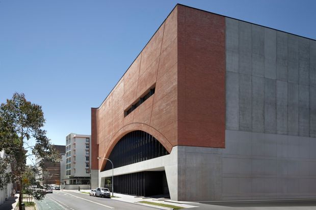 Street-level architectural photograph of a large urban building with a bold geometric massing. The facade combines extensive red brickwork with large smooth grey concrete panels, a tall curved arched opening filled with dark vertical glazing or louvers, and narrow horizontal slit windows. A wide street frontage and neighbouring mid-rise buildings reinforce the dense urban setting.
