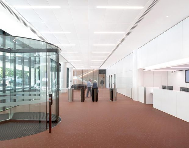 A bright, modern commercial interior showing a spacious reception or circulation area. The space features a large curved glass-enclosed stair or lift enclosure on the left, a white suspended-panel ceiling with linear lighting, white wall panelling and reception/service joinery on the right, glazed partitions at the rear, and a muted terracotta patterned floor. The composition has a clean, institutional corporate character with extensive glazing and open circulation space.