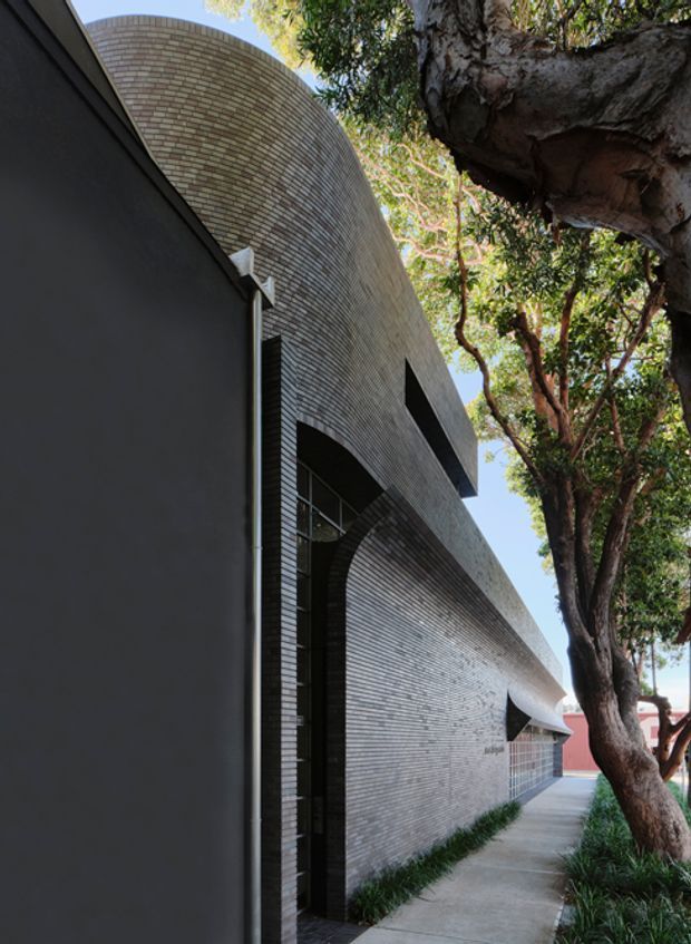 Exterior architectural view of a contemporary building along a narrow landscaped walkway. The facade is composed of pale, fine-grained brickwork with a dramatically curved roofline and recessed arched openings, contrasted by a dark smooth wall and tall dark-framed glazing. Mature trees closely frame the building and a red building is visible in the distance.