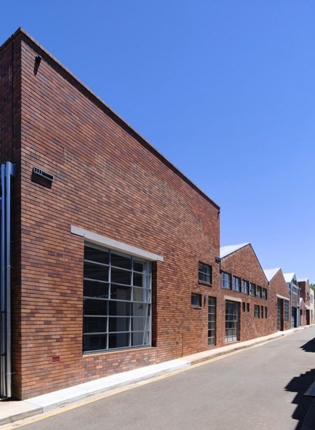 Exterior view of a long, low-rise brick building lining a narrow paved lane. The facade features dark red and brown stretcher-bond brickwork, large industrial-style multi-pane windows, smaller horizontal windows, exposed downpipes, and repeated pitched roof forms extending into the distance. Clear blue sky and strong sunlight emphasize the crisp urban-industrial character.