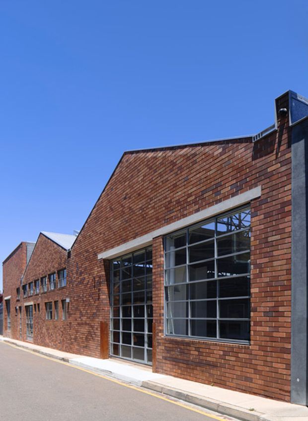 Exterior view of a low-rise commercial or industrial building with extensive red brick masonry, sawtooth or pitched roof forms, and large dark-framed multi-pane industrial windows and doors. The building fronts a paved street under a clear blue sky.
