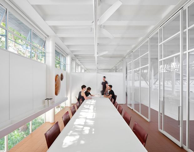 Bright, elongated contemporary commercial meeting or collaborative workspace with a long white conference table, burgundy upholstered chairs, white built-in wall storage, extensive glazed partitions, and large windows. The white exposed ceiling structure and ceiling fans create an airy, light-filled office environment, with warm terracotta-toned flooring and greenery visible outside.