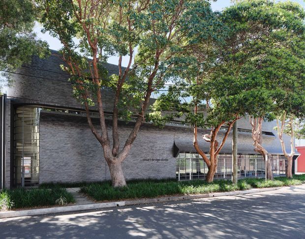 Street-facing view of a low-rise contemporary commercial or institutional building partially screened by mature trees. The facade uses dark grey masonry or brickwork, extensive glazing, and a curved glazed entrance volume, with landscaped planting along the footpath and a red-clad section visible at the right.