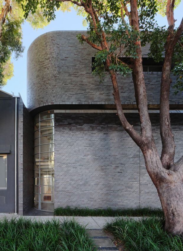 Street-facing view of a contemporary multi-storey building with a strongly rounded corner clad in narrow grey masonry units. A tall curved glazed window wraps the lower corner, while dark recessed window openings and horizontal bands define the facade. Mature trees and low ornamental planting partially screen the building, with a paved path leading to the entrance.
