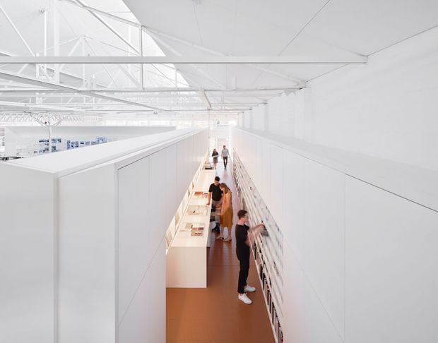 Bright, minimalist commercial interior showing a long elevated gallery or exhibition circulation space. White partitions, walls, ceiling, and exposed roof trusses create a highly uniform setting, with a warm terracotta-toned floor, display counters, railing, and several visitors. The space appears to be inside a large, airy hall with high-level glazing or skylights and strong linear perspective.