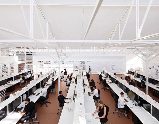 A bright, open-plan commercial design studio or architectural office with rows of long white workstations, desktop monitors, drafting and presentation materials, and many people working. The space has a high white ceiling with exposed structural trusses and beams, extensive white built-in shelving and display boards, and a warm brown floor. The overall appearance is spacious, collaborative, and highly illuminated.