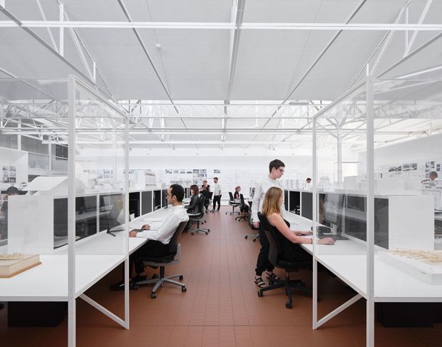 Bright open-plan commercial office or studio with rows of white workstations, translucent partition panels, rolling office chairs, and several people working. The space has a high white exposed structural ceiling with visible beams and services, extensive overhead glazing or skylight-like illumination, white steel columns and frames, and a terracotta-toned tiled floor. The layout feels airy, functional, and industrial-modern.