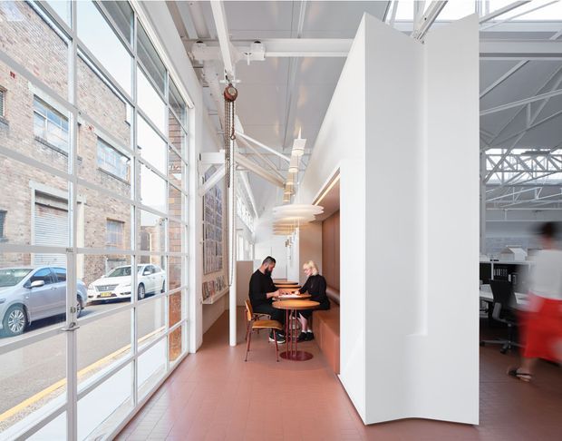 Bright adaptive commercial interior in a converted industrial building, featuring a tall glazed facade, exposed white-painted structural beams and services, terracotta tiled flooring, and a café-style seating nook integrated into a white and timber-lined partition. An open office or communal workspace is visible beyond the partition. The space has a high ceiling, strong natural light, and a contemporary industrial character.