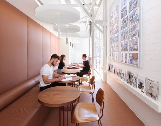 Bright, elongated commercial café or hospitality interior with a warm brown banquette along a terracotta-toned wall, round timber tables, and sculptural wooden chairs. A long white-painted brick wall displays framed architectural imagery and magazines, while large windows provide abundant daylight. The ceiling features exposed white structural framing, linear skylight-like glazing, and suspended circular acoustic panels with pendant lights. The space has a refined contemporary design with warm timber and leather accents.