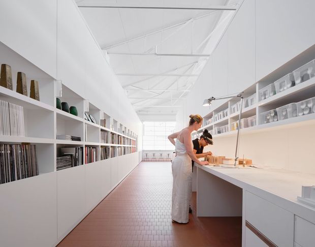 Bright, elongated interior workspace with a high white exposed-truss ceiling, full-length built-in white shelving and cabinetry, and a long pale work counter. Two people are working at the counter, while the shelving displays books, samples, and objects. The space has a clean, gallery-like, highly ordered appearance with warm reddish-brown tiled flooring and abundant daylight from the glazed end wall.