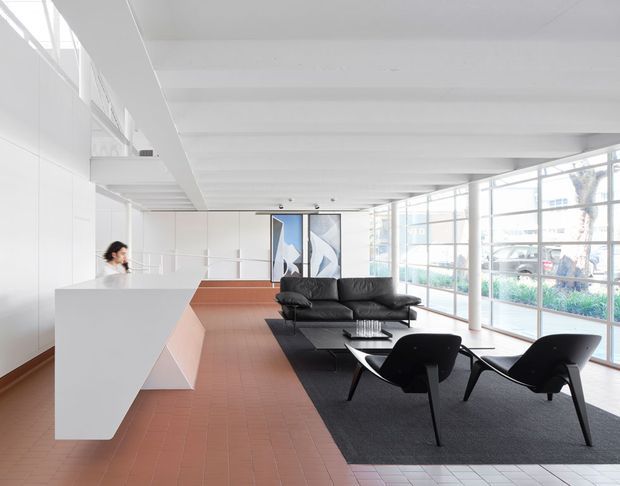 A bright, open-plan contemporary interior with a minimalist reception or kitchen-like white built-in counter on the left, a black leather sofa and sculptural black lounge chairs arranged on a dark rug, and extensive floor-to-ceiling glazing along the right wall. The space features a white exposed-beam ceiling, terracotta-colored tiled flooring, white wall panels, and abstract artwork. The overall composition is clean, spacious, and modern, with strong natural light.