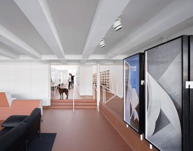 A bright, contemporary office or commercial interior with a long corridor-like lounge and gallery space. The room features exposed white-painted ceiling beams, track lighting, pale white partitions and glazing, terracotta-toned tiled flooring and lower wall surfaces, a black sofa in the foreground, and large monochrome framed artworks along the right wall. A glazed meeting or workspace area is visible beyond, with open shelving and a person in the distance.