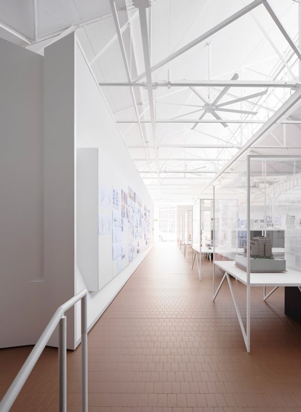 Bright, high-ceilinged architectural exhibition or studio interior with exposed white-painted roof trusses and services, extensive glass display partitions, pin-up boards, tables, and architectural models. The long circulation space has reddish-brown rectangular tile flooring, white walls, and abundant diffuse daylight, creating a clean gallery-like atmosphere.