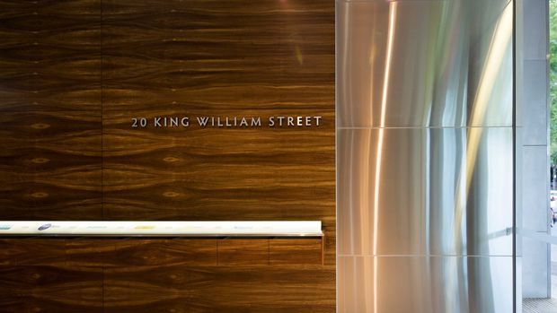 A refined commercial building lobby or reception area featuring a dark, richly grained timber feature wall with the text “20 King William Street,” a slim white reception counter or ledge, and a tall polished metallic cylindrical column beside floor-to-ceiling glazing. The composition is architectural and minimalist, with reflective surfaces and natural daylight.