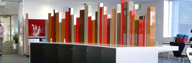 A contemporary commercial interior, likely a reception or waiting area, featuring a long black-and-white reception counter in the foreground and a striking sculptural screen behind it made from vertical timber-toned and red panels of varying heights. The space has white walls, polished light flooring, modern furniture, large windows, and bright, clean lighting.