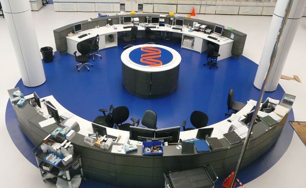 A circular, high-tech commercial control or operations room viewed from above, with curved workstations arranged around a central round console bearing a blue-and-orange emblem. Multiple computer monitors, telephones, office chairs, storage units, and equipment are arranged across a vivid blue floor, with large white structural columns surrounding the space.