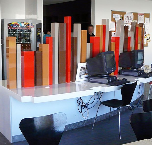 A contemporary commercial interior resembling a reception or service counter, with a long white counter displaying tall red, orange, grey, and wood-toned vertical panels. Computer monitors and keyboards sit on the counter, with black visitor chairs in front. A vending machine and bulletin board are visible in the background.
