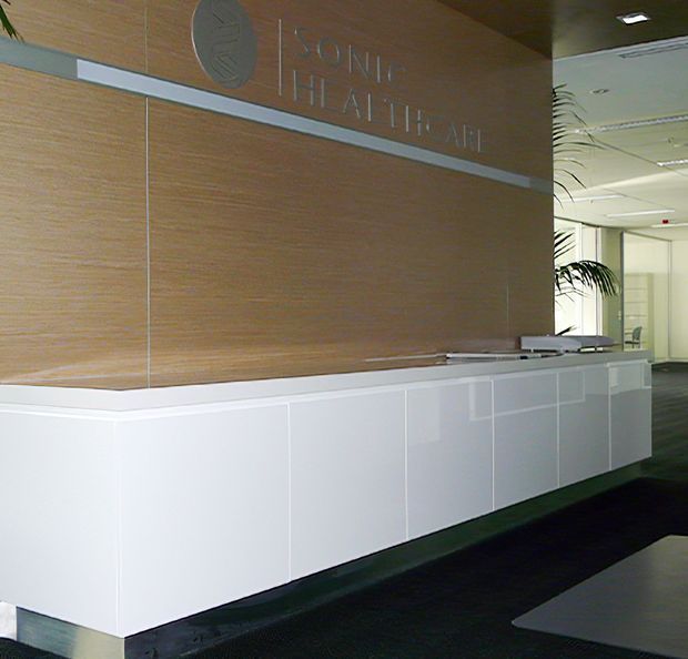 A modern corporate reception area featuring a long white built-in reception desk with integrated panel detailing, set against a warm wood-panelled feature wall with subtle raised signage. A corridor with glazed partitions and indoor planting is visible beyond the desk. The space has a clean, restrained commercial interior with dark flooring and recessed ceiling lighting.