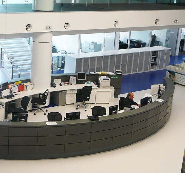 A spacious modern commercial reception or service desk area viewed from above, featuring a large curved dark-grey counter with multiple computer workstations, office chairs, printers, and staff desks. The space has white flooring, exposed cylindrical columns, extensive internal glazing, a blue accent floor area, and an upper-level mezzanine or balcony with a glass balustrade.