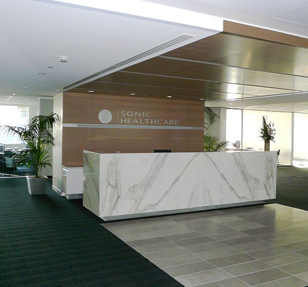 A bright healthcare reception lobby featuring a large white marble-look reception desk, warm timber wall panelling with Sonic Healthcare signage, a timber-lined ceiling feature, dark carpet, grey tiled flooring, indoor plants, and expansive glazing.